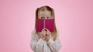 Eğitim, okul konseptine geri dönelim. Oynayan küçük kız yüzünü defterle kapatıyor ve dışarı bakıyor, kameraya bakıyor, pembe stüdyo arka planı, ağır çekim