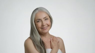 Vücuttan vücuda. Üst düzey gri saçlı, şımarık krem kalbini omzuna sürten, kameraya gülümseyen, beyaz stüdyo arka planında havluya sarılı poz veren, yavaş çekim yapan bir kadın.