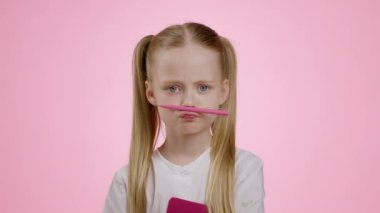 Bıyık şakası. Burun ve dudaklar arasında kalem tutan, pembe stüdyo arka planında, yavaş çekim yapan, pozitif küçük okul kızının yakın portresi.