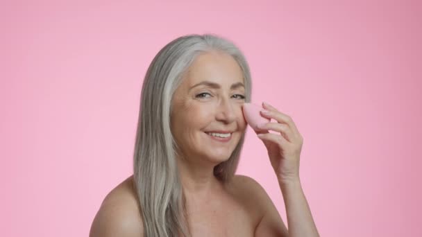 美人の秘密 特殊メイクスポンジでコンシーラーを適用幸せな白髪のシニア女性のスタジオポートレート カメラに笑顔 ピンクの背景 スローモーション — ストック動画