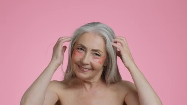 朝の美しさの儀式 スタジオポートレートの屈託のないシニア女性とともに抗浮腫水腫パッチ下目で遊んで彼女の灰色の髪 カメラに笑みを浮かべて ピンクの背景 スローモーション — ストック動画