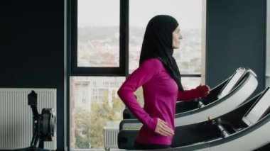 Günlük hayatta faaliyet. Spor salonundaki koşu bandında koşan geleneksel tesettürlü Müslüman kadının yan görünüş portresi. İzleme çekimi, yavaş çekim.