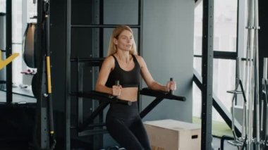 Karın kası egzersizi. Sportif orta yaşlı kadın sporcu spor salonundaki spor barlarında egzersiz yapıyor, bacaklarını kaldırıyor, vücut gücü antrenmanı yapıyor, yavaş çekim, serbest alan.