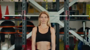 Pazı egzersizi. Pozitif orta yaşlı kadının portresi. Dambılları kaldırıyor, modern spor salonunda el kaslarını çalıştırıyor, kameraya gülümsüyor, yavaş çekim.