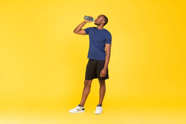 运动健美的黑人男子从健康瓶喝水站在黄色工作室的背景 健身训练中的男性水合物 锻炼身体和健康的水合物概念 全长铅球 — 图库照片