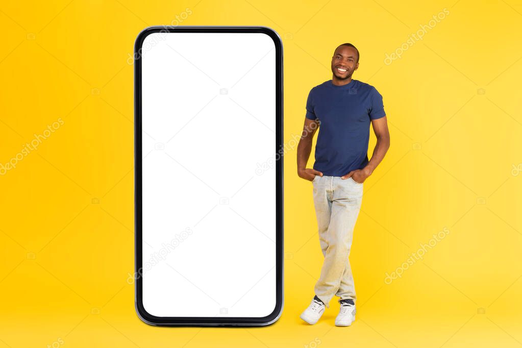 Happy Black Guy Standing Near Big Cellphone Blank Screen Posing Smiling To Camera In Studio Over Yellow Background. New Mobile Application Concept. Full Length Shot, Mockup