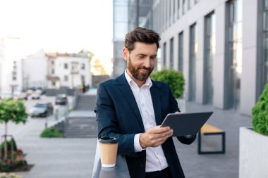 Kendine güveni tam, takım elbiseli beyaz bir adam modern ofis binasının balkonunda tablete yazı yazıyor. Başarılı bir iş ve kahve molası, iş adamının sosyal uzaklık nedeniyle video görüşmesi var.