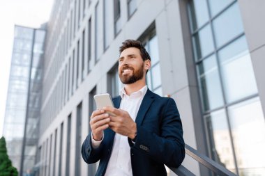 Kendine güveni tam, takım elbiseli beyaz bir adam dışarıda akıllı telefondan yazı yazıyor ve modern ofis binasının yakınındaki boş alana bakıyor. Reklam ve iş teklifi, iş adamı ticareti uygulaması