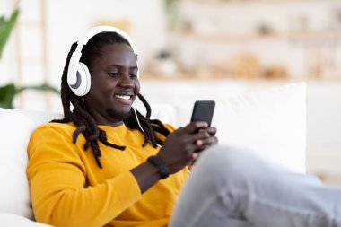 Mutlu siyahi adam kulaklıkla müzik dinliyor ve evde akıllı telefon kullanıyor, cep telefonuyla koltukta dinlenirken gülümseyen Afrikalı Amerikalı genç adam, boş alanla yakın çekim yapıyor.