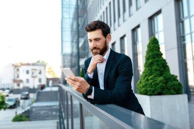 Dalgın gülümseyen, kendine güveni tam, takım elbiseli beyaz adam modern ofis binasının balkonunda akıllı telefondan izliyor. İş adamı cihaz, iş uygulaması, video çağrısı ve çalışma kullanır