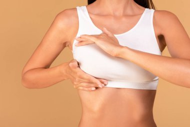 Göğüslerini gösterme konsepti. Beyaz sütyenli genç bir kadın kanser önleme için kontrol yapıyor, bej renkli stüdyo geçmişi, yakın plan, kırpılmış