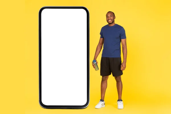 Sporty Black Male Posing Near Mobile Phone With Large Empty Screen Holding Water Bottle Standing In Studio On Yellow Background. Great Fitness Workout Application Concept. Full Length, Mockup