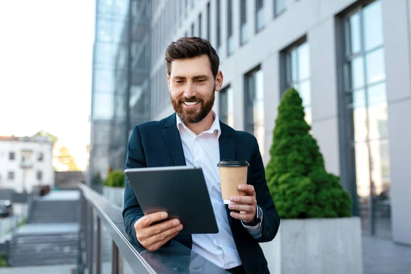 Smiling confident young caucasian guy with beard in suit has video call at tablet, hold cup of takeaway coffee outdoors on balcony of modern office building. Business, new normal for work with gadget