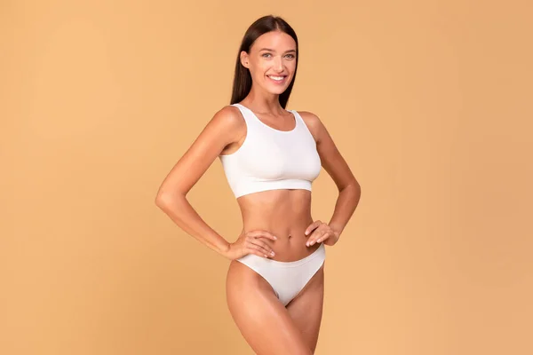 Body slimming concept. Happy lady with slim figure in white underwear smiling at camera, fit woman with flat abdomen isolated over beige studio background