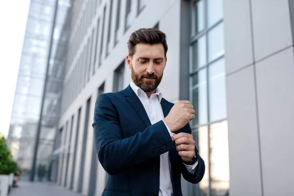 Smiling confident calm attractive young caucasian male ceo manager with beard in suit near modern office building, outdoor, copy space. Successful business, businessman in city, ad, boss and career
