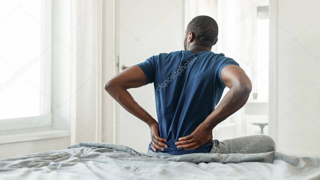 Back View Of African American Man Having Pain Touching Lower Back Sitting On Bed In Modern Bedroom Indoors. Uncomfortable Bed And Spine Health Problem Concept. Panorama
