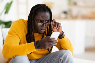 Kötü Vizyon. Gözlüklü genç siyah adam evde akıllı telefon ekranına bakıyor, cep telefonundan mesaj okumaya çalışan Afrikalı Amerikalı adam, acı çeken ya da görme bozukluğu olan,