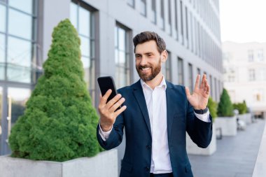 Takım elbiseli, sakallı, kendine güveni tam, yakışıklı beyaz bir menajer ofis binasının yakınında video görüşmesi yapıyor. İş adamı ve açık hava konferansı, başarılı iş ve yeni normal