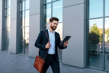 Sakallı, takım elbiseli, tabletle sohbet eden yakışıklı, kendine güvenen beyaz adamın sokaktaki modern ofis binasında çalışmasına sevindim. İş adamı iş, finans ve ticaret için uygulama kullanıyor, uzaktan görüşme