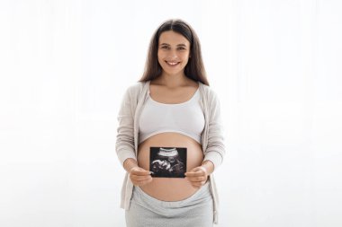 Bebeğimin ilk portresi. İç çamaşırlı, güzel, uzun saçlı hamile bir kadın ultrason görüntüsü gösteriyor ve kameraya gülümsüyor. Beyaz arka planda duruyor.