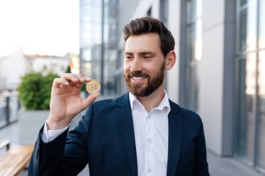 Takım elbiseli, kendine güveni tam beyaz bir adam Bitcoin madeni parayı modern ofis binasının yakınında gösteriyor. Başarılı çevrimiçi işler, iş adamı kar elde etmek için kripto para birimiyle çalışıyor.