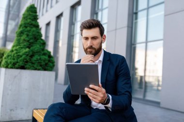Takımlı sakallı, düşünceli, çekici beyaz bir erkek tabletten haber okuyor, modern ofis binasının yanındaki bankta oturuyor. İş adamı planlama stratejisi, yeni normal ve uygulama