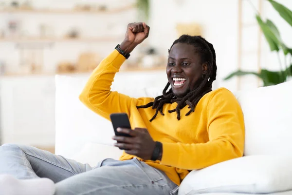 自宅で成功を祝うスマートフォンを持つ楽しいアフリカ系アメリカ人の男の肖像 興奮とリビングルームでソファに座って 電話画面を見て幸せな若い黒人男性 スペースをコピー — ストック写真