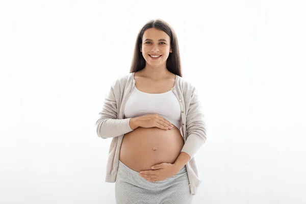 快適な家庭用で陽気なかなり若い妊婦は 彼女の大きな腹を示し カメラで笑みを浮かべて 美しい女性は赤ちゃんを待って 白い背景 コピースペース 妊娠概念 — ストック写真