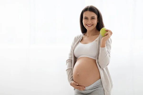 Cheerful beautiful long-haired young pregnant lady in homewear holding fresh green apple, smiling at camera, white background, panorama with copy space. Healthy diet during pregnancy