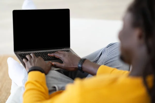 Young Black Man Typing On Laptop With Blank Screen While Sitting On Couch At Home, Unrecognizable African American Male Using Computer For Remote Work Or Online Education, Over Shoulder View, Mockup