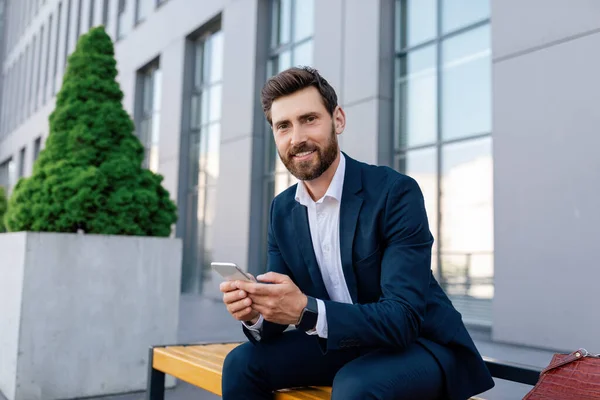 Successful business and health care. Happy confident young caucasian guy with beard in suit typing on smartphone near modern office building. Application for conference outdoor, gadget and new normal