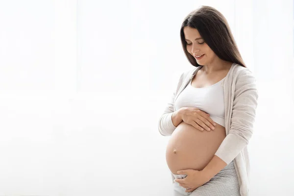 快適なホームウェアで幸せな若い妊婦の自宅でウィンドウの横に立って 彼女の大きなおなかを見て 赤ちゃんを待って コピースペースとパノラマ 妊娠中や母親の考え方 — ストック写真