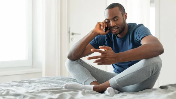 自宅のモダンなベッドルームでベッドの上に座って通信携帯電話で話す黒人男性 スマートフォンの屋内で話す男 現代のコミュニケーション パノラマ — ストック写真