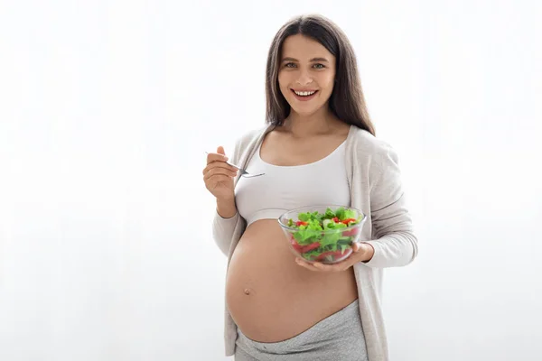 Nutrition and healthy diet during pregnancy concept. Happy pregnant woman holding bowl with fresh vegetable salad and fork, smiling at camera, white background, free space