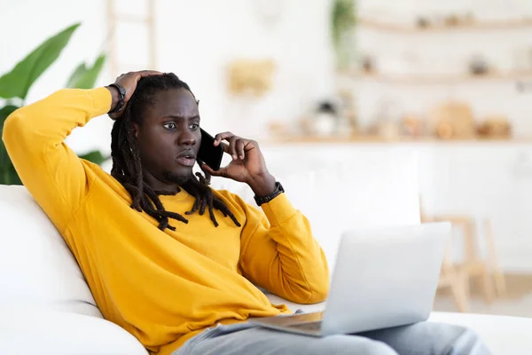 Internet Scam. Shocked Black Man Looking At Laptop Screen And Talking On Cellphone, Confused Young African American Male Having Problems With Computer, Sitting On Couch At Home, Copy Space