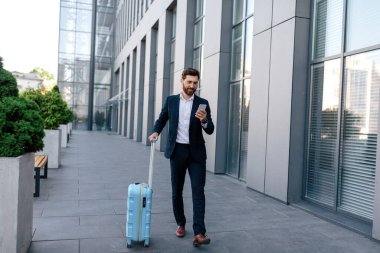 Bavullu, sakallı, kendine güveni tam, yakışıklı beyaz bir erkek modern ofis binasının yakınında, açık havada, akıllı telefon ile sohbet ediyor. İş gezisi, iş adamı uygulama haritasını aygıtta kullan