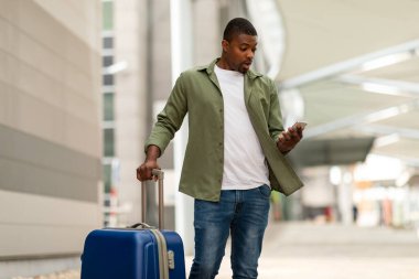 Şok olmuş siyah turist, açık hava limanındaki seyahat çantasıyla telefonuyla Negatif Mesaj okuyor. Mobil İletişim Sorunları Konsepti