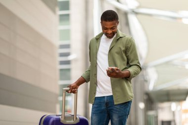 Mutlu Siyah Turist Seyahat Başvurusu ile Havalimanı 'nda bavulla ayakta duran telefonu kullanıyor. Guy biletlerini internetten ayırtıyor. Turizm İçin Gadget ve Mobil Hizmetler