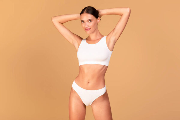 Портрет счастливой молодой женщины в женском белье, позирующей на фоне бежевой студии, поднимающей руки вверх. Женщина с плоским животом в белом нижнем белье верхней бюстгальтер и трусики