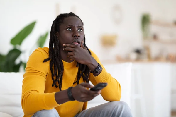 手握遥控器坐在沙发上坐在家里的惊心动魄的黑人男子的画像 恐怖的非洲裔美国男性观看电视节目时对惊心动魄的内容的反应 复制空间 — 图库照片