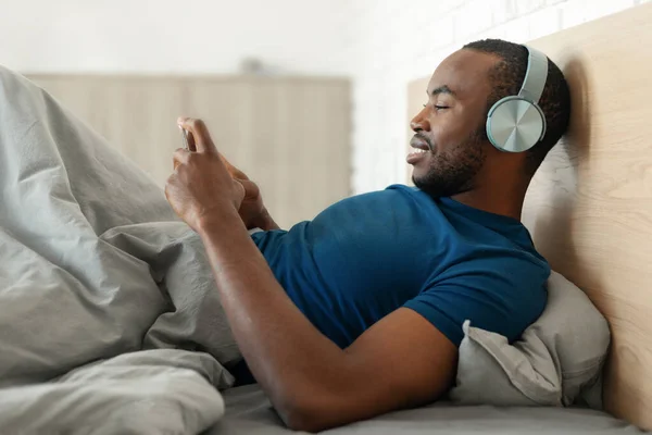 携帯電話を身に着けている携帯電話を使用して幸せなアフリカ系アメリカ人男性は 現代の寝室の屋内で居心地の良いベッドでリラックスして音楽をオンラインで聴いています サイドビューショット — ストック写真