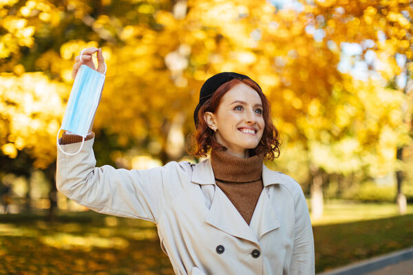 Smiling caucasian millennial red-haired lady in raincoat and beret takes off and throws out protective mask in park in autumn with yellow leaves. Freedom and health care, end of covid-19 quarantine