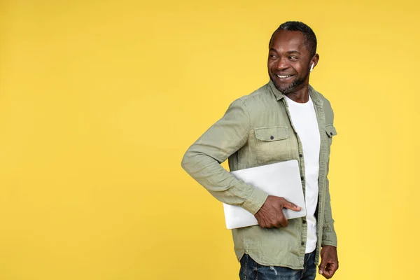Smiling confident mature black male in casual, wireless headphones with laptop looking back at empty space, isolated on yellow background, studio. Great ad and offer, technology for work and business