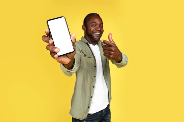 オンライン勝利へのカジュアルな喜びで笑顔の大人の黒人男性は 黄色の背景 スタジオに隔離された空白の画面でスマートフォンで指を表示します 人々の感情の成功と勝利 アプリ 広告やオファー — ストック写真