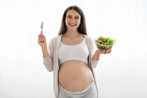 幸せそうな魅力的な長髪の若い妊娠中の女性ホームウェアで新鮮な野菜サラダとフォークとガラスボウルを保持 陽気にカメラで笑顔 白の背景 コピースペース — ストック写真