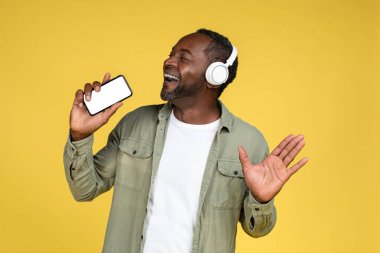 Sıradan, kablosuz kulaklıklı neşeli, olgun Afrikalı Amerikalı erkek hayali mikrofonda şarkı söylüyor ve sarı arka planda, stüdyoda izole edilmiş akıllı telefonuyla şarkı söylemekten hoşlanıyor. Ses uygulaması olan bir süperstar