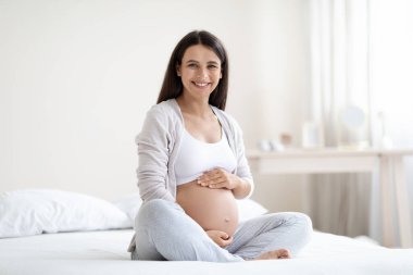 Ev elbiseli mutlu, genç, hamile bir kadın evde yatağında oturuyor, koca göbeğine sarılıyor ve neşeli bir şekilde kameraya gülümsüyor, beyaz yatak odasının içi, fotokopi odası. Hamilelik, annelik kavramı