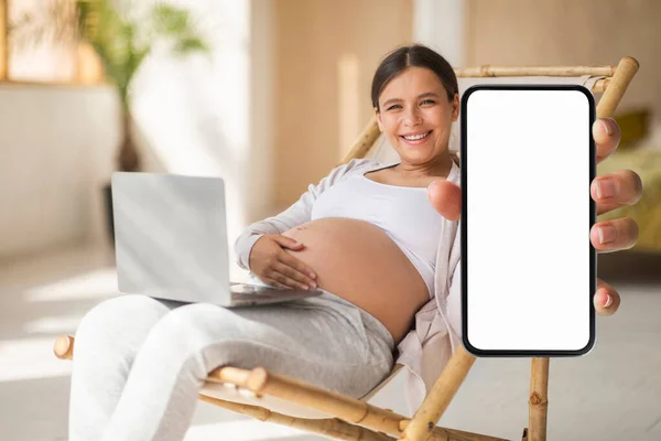 Pregnancy Tracker App. Pregnant woman showing blank smartphone while resting in chair with laptop, happy expectant female demonstrating modern cellphone with copy space for design, mockup