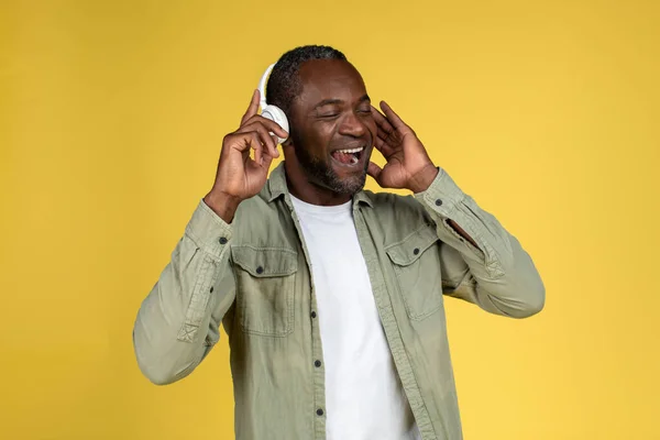 Satisfied mature black man in casual with wireless headphones with closed eyes enjoy music, singing alone, have fun, isolated on yellow background, studio. Entertainment with audio app and lifestyle