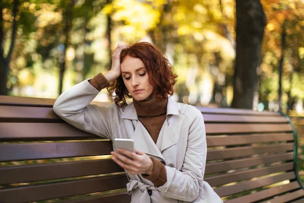 Lavičce Sedí Nešťastná Běloška Pláštěnce Podzim Dívá Smartphone Parku Žlutými — Stock fotografie